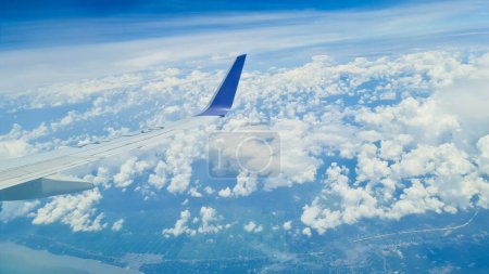 Wing of an airplane with a blue tip with beautiful blue sky and white cloud as background and the land and river in the distance