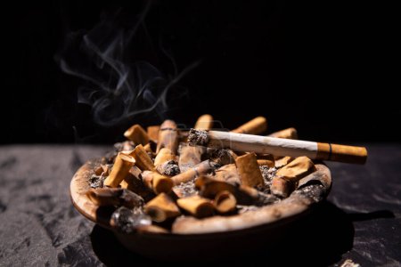 Photo for Cigarettes burning with light smoke in ashtray with black background - Royalty Free Image