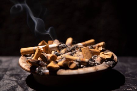 Photo for Smoldering cigarette in ashtray full of cigarette stubs on against black background - Royalty Free Image