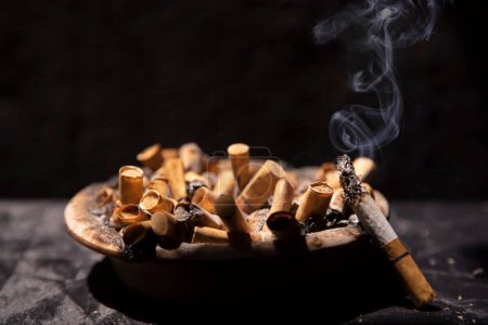 Photo for Smoldering cigarette with smoke in ashtray full of cigarette stubs - Royalty Free Image