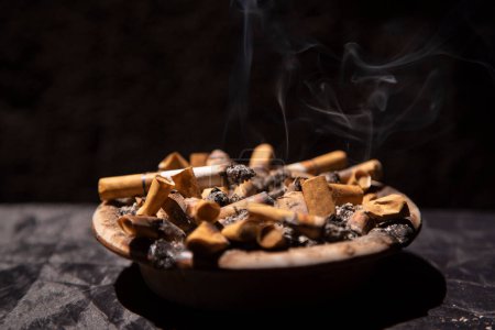 Photo for Cigarette in ashtray full of cigarette stubs on against black background - Royalty Free Image