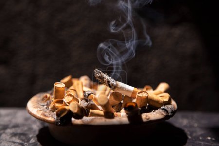 Photo for Smoky cigarette in ashtray full of stubs - Royalty Free Image
