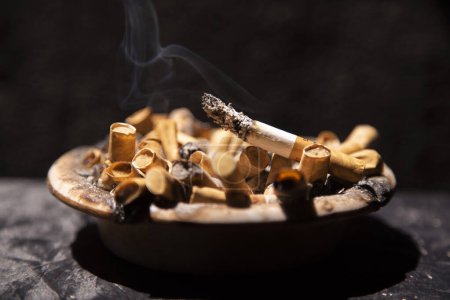 Photo for Burning cigarette with smoke in ashtray full of stubs - Royalty Free Image