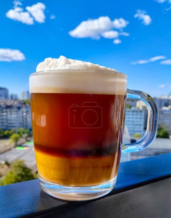 Photo for A glass cup of cappuccino with airy foam stands on the frame of a balcony against the blue sky. In the background there is a city landscape without focus. Vertical photo. - Royalty Free Image