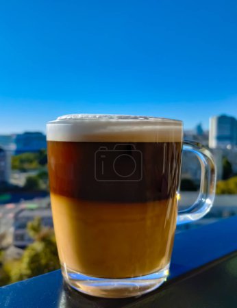 Photo for A glass cup of cappuccino with airy foam stands on the frame of a balcony against the blue sky. In the background there is a city landscape without focus. Vertical photo. - Royalty Free Image