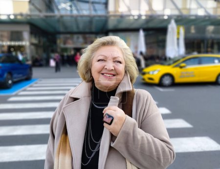 Porträt einer schönen reifen, glücklichen Seniorin in beigem Mantel und riesigem Ring, die in der Nähe eines Einkaufszentrums steht