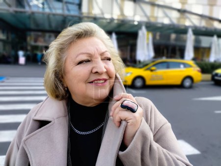 Portrait of a beautiful mature happy senior woman in beige coat and huge ring standing near shopping mall