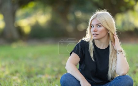 Eine junge Frau mit Zahnspange sitzt im Gras, die Hand auf dem Kopf, während sie in einem Park lächelt.