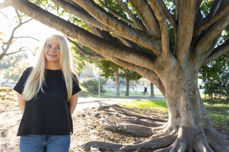Eine junge Frau mit Zahnspange steht neben einem massiven Baum in einem Park und lächelt in die Kamera.