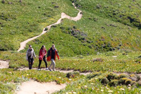 Three friends are hiking up a trail in the mountains. They are surrounded by towering peaks and lush greenery as they make their way up the path together.