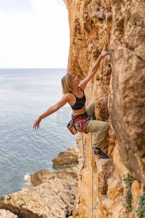 Photo for A woman is climbing a rock wall with a rope. The scene is set against a backdrop of the ocean - Royalty Free Image