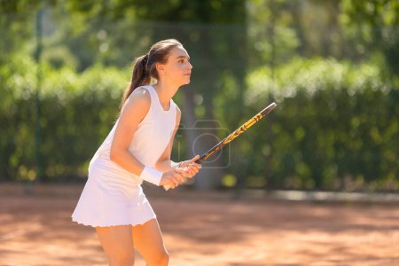 A woman in a white dress is playing tennis on a court. She is holding a tennis racket and is ready to hit the ball
