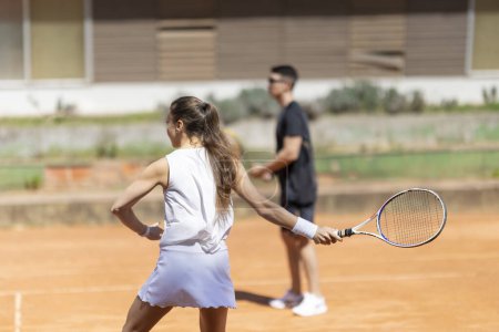 A woman is playing tennis with a man watching her. The woman is wearing a white shirt and skirt