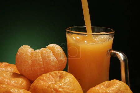 Obst, eine geschälte und andere ungeschälte Orangen mit Orangensaft im Glas vor grün-schwarzem Hintergrund