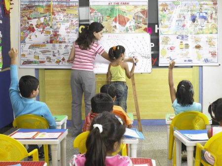 Photo for South Asian Indian girl writing on a board and teacher helping her in nursery school - Royalty Free Image