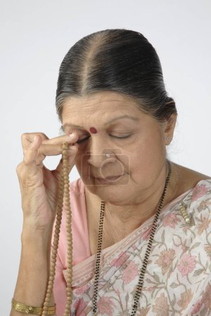 Alte Dame betet mit Rosenkranz oder Japmala in der rechten Hand, die das rechte Auge berührt 