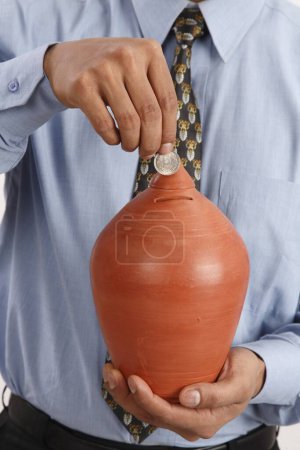 Foto de Empresario sosteniendo pequeña olla de tierra de ahorro en una mano e insertando a través de ranura moneda de cinco rupias MR703T - Imagen libre de derechos