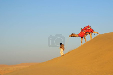 Foto de Camello con propietario caminando por la pendiente de dunas de arena blanda en Sam; Sam; Jaisalmer; Rajastán; India - Imagen libre de derechos