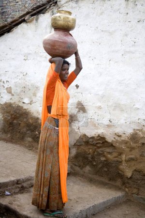 Foto de Mujer rural llevando agua en maceta de barro sobre su cabeza de pie en el carril de la aldea; pueblo semi urbano Dilwara; Udaipur; Rajastán; India - Imagen libre de derechos