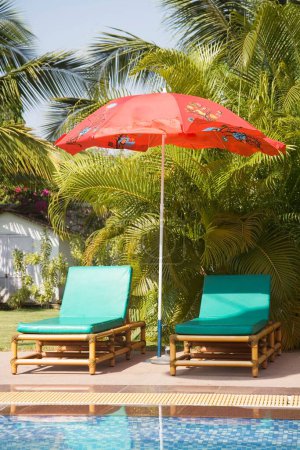 Téléchargez les photos : Parapluie de plage unique de couleur rouge près de la piscine d'eau bleue palmiers verts et ciel bleu ; Palolem plage ; Goa ; Inde - en image libre de droit