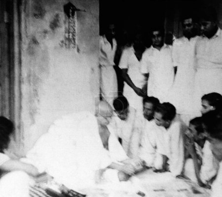 Foto de Mahatma Gandhi hablando con la gente mientras revisaba los periódicos durante su visita a la zona afectada por los disturbios de Noakhali, Bengala Oriental, noviembre de 1946, India - Imagen libre de derechos