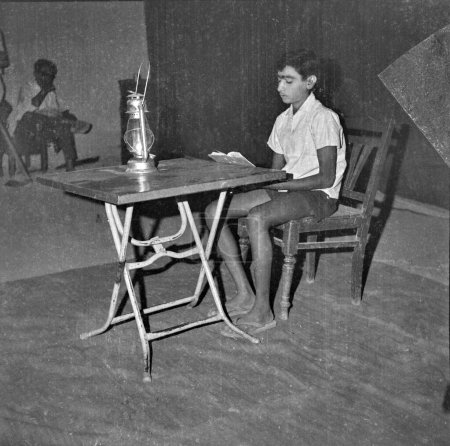 Foto de Viejo vintage 1900s foto en blanco y negro niño indio estudiar kerosene lámpara linterna luz aluminio mesa sentado silla de madera India 1940 - Imagen libre de derechos