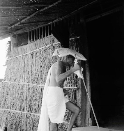 Téléchargez les photos : Mahatma Gandhi, portant un sac de boue sur sa tête, entrant dans une cabane à l'ashram de Sevagram, 1941 - en image libre de droit