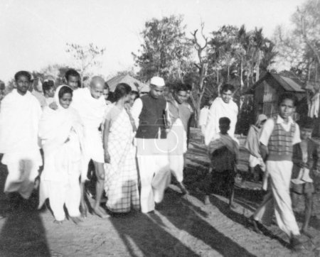 Foto de Mahatma Gandhi, con el apoyo de Amtus Salam y Abha Gandhi, caminando con Sachin Mitra y otros a través de campos en las zonas afectadas por disturbios en Noakhali Bengala Oriental, noviembre 1946, India - Imagen libre de derechos
