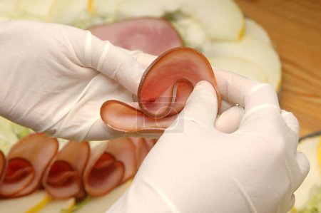 Non vegetarian food , hand with white rubber gloves , making rose flower shape of smoked ham round slices to arrange in cold platter with smoked ham and honey melon decorated platter