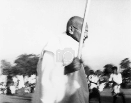 Foto de Mahatma Gandhi caminando con un gran palo de bambú a través de la zona afectada por disturbios musulmanes hindúes en Noakhali Bengala Oriental, noviembre 1946, India - Imagen libre de derechos