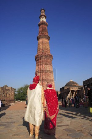 Téléchargez les photos : Couple de mariage américain en costume indien devant Qutb Minar construit en 1311 tour de grès rouge, art indo-musulman, sultanat de Delhi, Delhi, Inde Site du patrimoine mondial de l'UNESCO - en image libre de droit