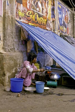 Foto de Un habitante del pavimento cocinando su comida en un refugio de cambio rodeado de carteles de películas de Bollywood, Mumbai Bombay, Maharashtra, India - Imagen libre de derechos