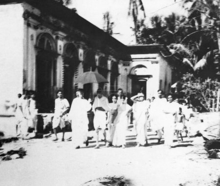 Foto de Mahatma Gandhi, caminando con Abha Gandhi, Sushila Nayar, Sushila Pai y otros en frente de un edificio en el área afectada por disturbios musulmanes hindúes en Noakhali Bengala Oriental, noviembre 1946, India - Imagen libre de derechos