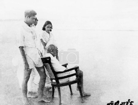 Téléchargez les photos : Mahatma Gandhi, assis sur une chaise Juhu Beach, Mumbai, avec des gens autour de lui, mai 1944, Inde - en image libre de droit