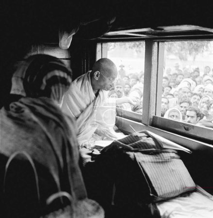 Foto de Mahatma Gandhi, sentado en un compartimiento del tren, escribiendo con su mano derecha y recogiendo donaciones para el Fondo Harijan con la mano izquierda, 1945 - Imagen libre de derechos