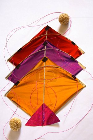 Colourful kites with thread and sweet tilgur or til gul sesame laddoos for Makara Sankranti festival on white background