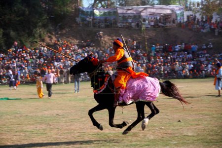 Foto de Nihang o guerrero sij llevando lanza y rifle realizando acrobacias a caballo durante el festival Hola Mohalla en el sahib de Anandpur en el distrito de Rupnagar, Punjab, India - Imagen libre de derechos