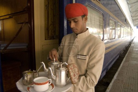 Téléchargez les photos : Rajasthani préposé tenant plateau de boisson debout à l'extérieur du patrimoine sur roues Voyage dans le train touristique de luxe passé ; Rajasthan ; Inde - en image libre de droit