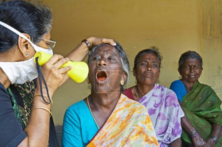 Foto de Trabajador sanitario de la ONG Chinmaya Organización de Desarrollo Rural CORD inspeccionando garganta de paciente rural esperando en el centro de salud, Siruvani, Karnataka, India - Imagen libre de derechos
