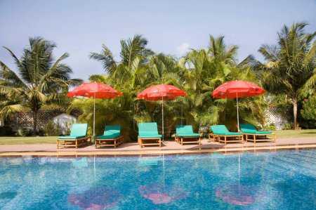 Photo for Three red color beach umbrella near the swimming pool blue water green palm trees and blue sky ; Palolem beach ; Goa ; India - Royalty Free Image