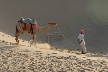 Foto de Hombre con camello caminando en dunas de arena; Jaisalmer; Rajastán; India - Imagen libre de derechos
