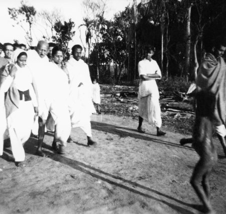 Téléchargez les photos : Mahatma Gandhi, soutenu par Amtus Salam, marchant avec d'autres à travers les champs dans les zones touchées par les émeutes au Noakhali Bengale oriental, Novembre 1946, Inde - en image libre de droit