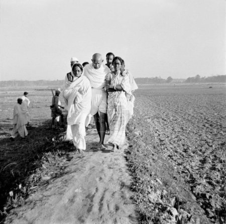 Foto de Mahatma Gandhi, con el apoyo de Amtus Salam y Abha Gandhi, caminando con Sachin Mitra y otros a través de campos en las zonas afectadas por disturbios en Noakhali Bengala Oriental, noviembre 1946, India - Imagen libre de derechos