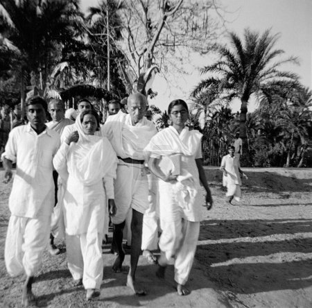 Téléchargez les photos : Mahatma Gandhi, soutenu par Amtus Salam et Manu Gandhi, marchant avec d'autres à travers les zones touchées par les émeutes à Noakhali, Bengale oriental, Novembre 1946, Inde - en image libre de droit