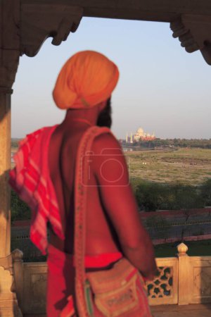 Téléchargez les photos : Sadhu observant Taj Mahal vue depuis le fort d'Agra construit au XVIe siècle par l'empereur moghol sur la rive ouest du fleuve Yamuna, Agra, Uttar Pradesh, Inde Site du patrimoine mondial de l'UNESCO - en image libre de droit