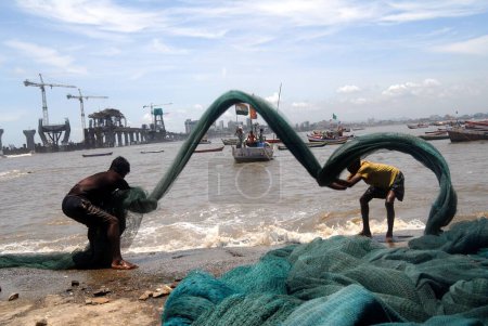 Photo for Fishermen clean their fishing nets in the backdrop of Bandra-Worli Sea Link at Worli Village in Bombay now Mumbai, Maharashtra, India - Royalty Free Image