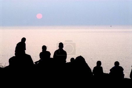 Téléchargez les photos : Touristes assis sur des rochers et regardant coucher de soleil à Harihareshwar Beach, Silhouette, Mer d'Arabie, Région de Konkan, District de Raigad, Maharashtra, Inde - en image libre de droit