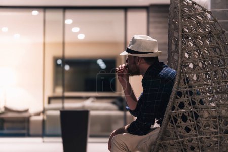Téléchargez les photos : Jeune homme fumant une cigarette électronique à la maison après une dure journée de travail. Concept de style de vie. - en image libre de droit
