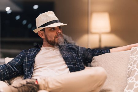 Junger Mann in legerer Kleidung raucht nach einem langen Arbeitstag zu Hause auf der Couch. Lifestyle-Konzept.