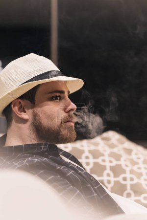 Raucher mit Hut im Wohnzimmer seines Hauses, der Rauch aus dem Mund bläst. Lifestyle-Konzept. Kopierraum.